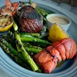 Grilled steak and lobster tail with asparagus, lemon wedges, and a creamy sauce on a platter.