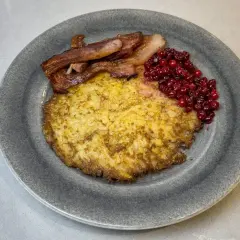 En gyllene raggmunk i ugn serveras med knaperstekt bacon och rårörda lingon på en grå tallrik.