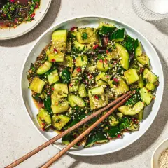 En skål med asiatisk gurksallad, toppad med sesamfrön och färska örter. En skål med dressing och ätpinnar ligger bredvid.