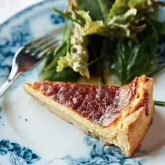 En bit gyllene ostpaj med olika ostar, serverad med en fräsch sallad på en vacker blåvit tallrik.