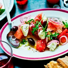 En uppfriskande melon och fetaostsallad med oliver, rödlök och ruccola, serverad med bröd och en drink.