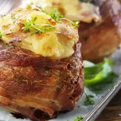Baked potatoes wrapped in bacon, topped with cheese and herbs, on a platter.