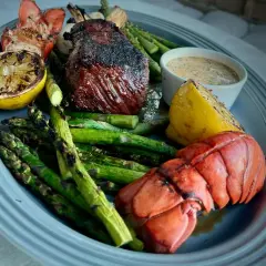 Grilled steak and lobster tail with asparagus, lemon wedges, and a creamy sauce on a platter.