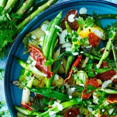 A close-up of a vibrant salad with grilled asparagus, potatoes, and crispy prosciutto, drizzled with a creamy dressing and garnished with fresh herbs.