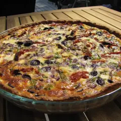 En gyllene paj utan skal, fylld med soltorkade tomater, oliver och lök, serveras på ett träbord.