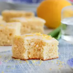 En bit saftig citronkaka i långpanna med glasyr och citronskal. En hel citron och en tulpan syns i bakgrunden.