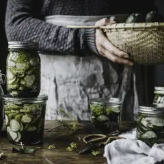 Burkar med inlagd gurka och dill, redo för ett gott saltgurka recept. En korg med färska gurkor i bakgrunden.