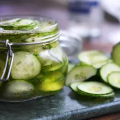 En glasburk med inlagd gurka, omgiven av skivade gurkor på en marmorplatta. Perfekt för sommaren!