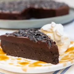 En saftig kladdkaka med lakritsströssel, serverad med vispad grädde och en skvätt sirap på en mönstrad tallrik.