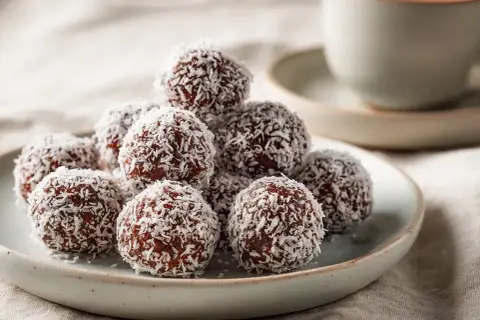 En hög med chokladbollar, rullade i kokosflingor, som ser ut att vara redo för ett chokladbollar recept. En kopp kaffe i bakgrunden.