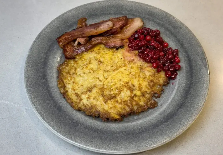 En gyllene raggmunk i ugn serveras med knaperstekt bacon och rårörda lingon på en grå tallrik.