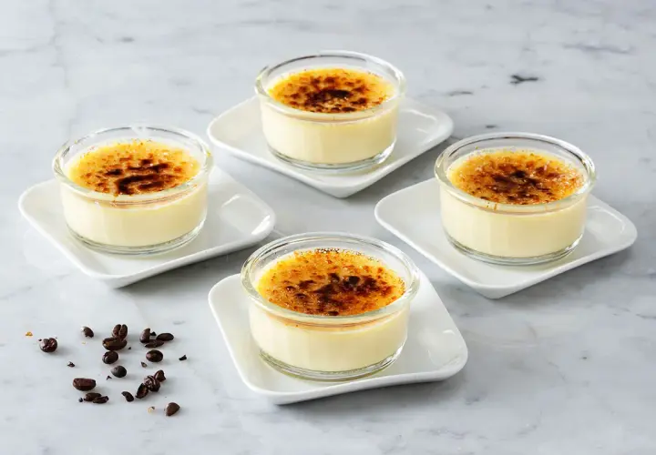 Fyra små glas med krämig creme brulee, toppad med karamelliserat socker. Kaffebönor ligger utspridda på marmorbordet.