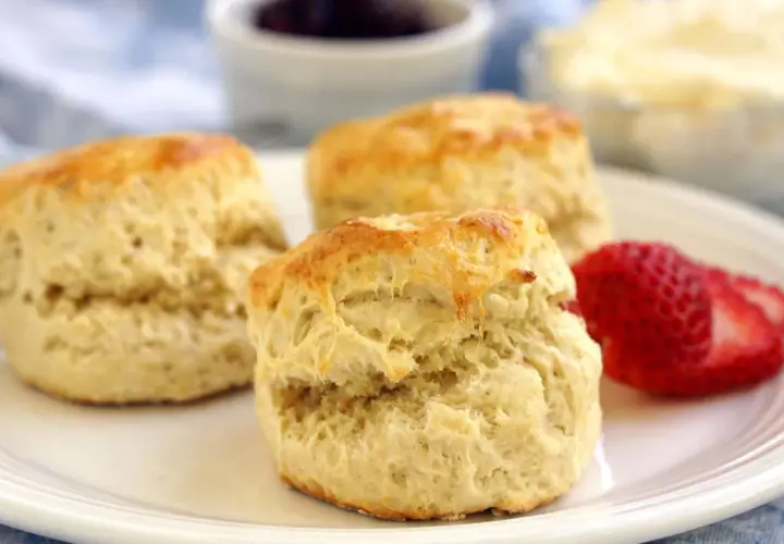 Tre gyllene scones på en vit tallrik, redo att avnjutas med sylt och grädde. Ett perfekt scones recept för fikat.