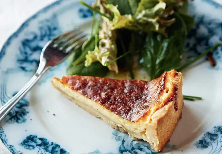 En bit gyllene ostpaj med olika ostar, serverad med en fräsch sallad på en vacker blåvit tallrik.