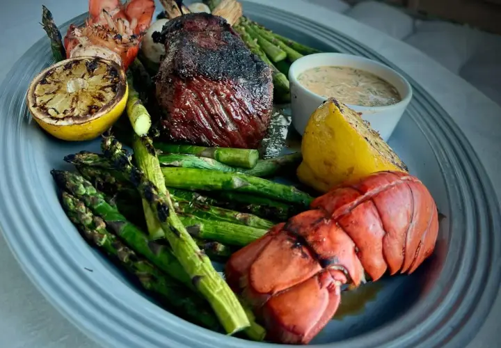 Grilled steak and lobster tail with asparagus, lemon wedges, and a creamy sauce on a platter.