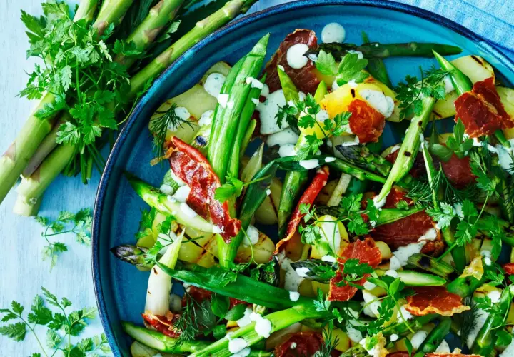 A close-up of a vibrant salad with grilled asparagus, potatoes, and crispy prosciutto, drizzled with a creamy dressing and garnished with fresh herbs.