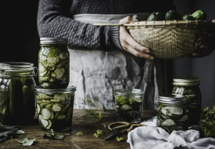 Burkar med inlagd gurka och dill, redo för ett gott saltgurka recept. En korg med färska gurkor i bakgrunden.