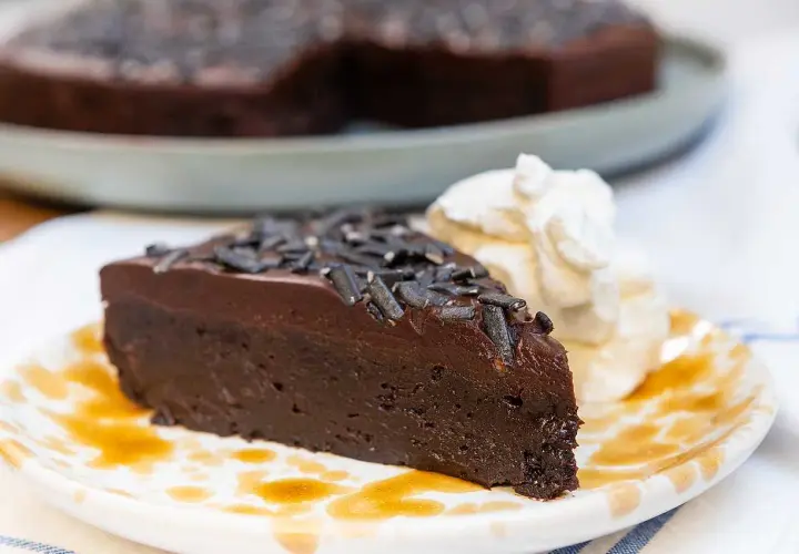 En saftig kladdkaka med lakritsströssel, serverad med vispad grädde och en skvätt sirap på en mönstrad tallrik.