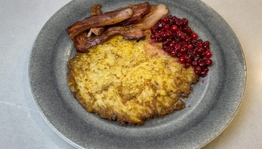En gyllene raggmunk i ugn serveras med knaperstekt bacon och rårörda lingon på en grå tallrik.