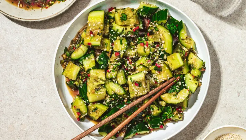 En skål med asiatisk gurksallad, toppad med sesamfrön och färska örter. En skål med dressing och ätpinnar ligger bredvid.