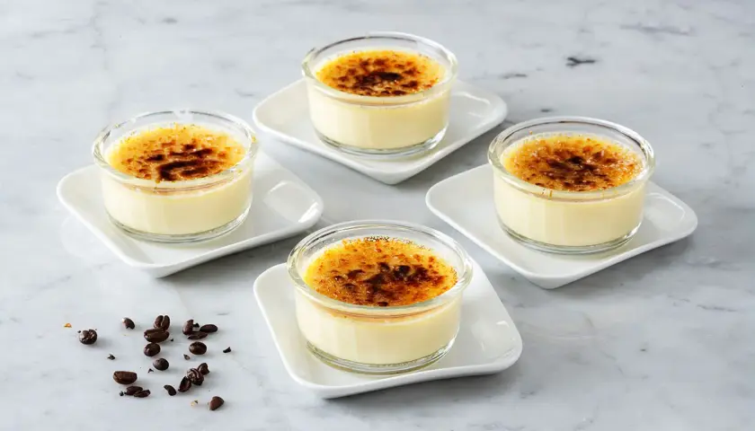 Fyra små glas med krämig creme brulee, toppad med karamelliserat socker. Kaffebönor ligger utspridda på marmorbordet.