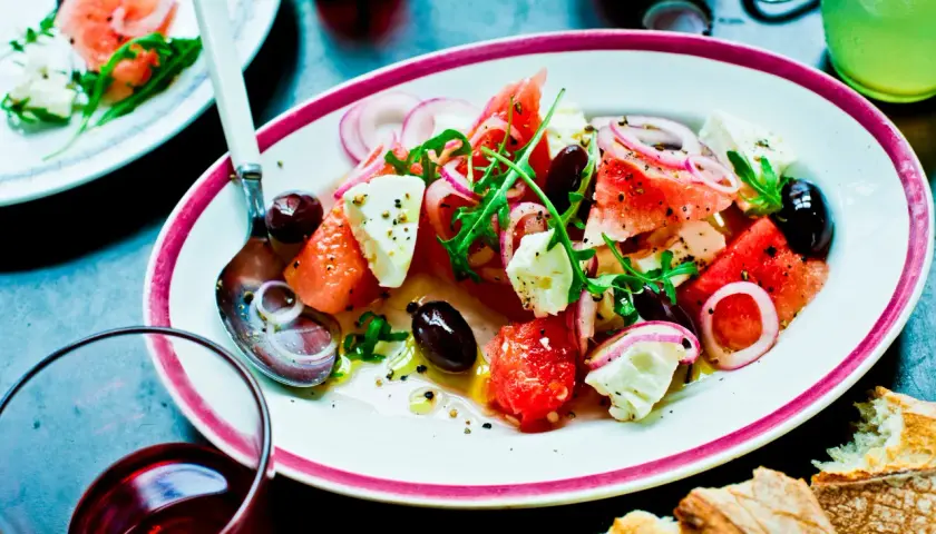 En uppfriskande melon och fetaostsallad med oliver, rödlök och ruccola, serverad med bröd och en drink.