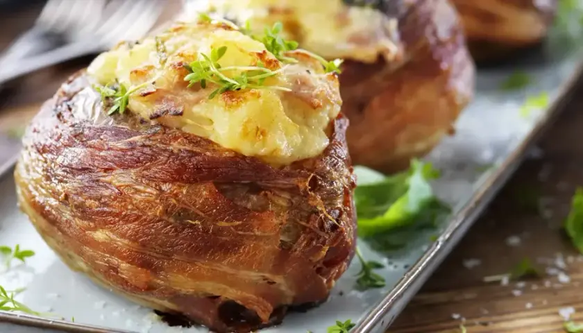 Baked potatoes wrapped in bacon, topped with cheese and herbs, on a platter.