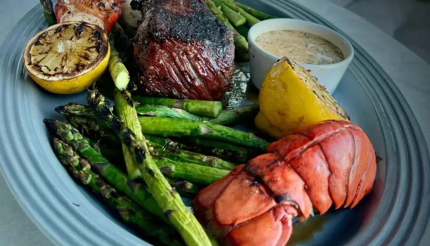 Grilled steak and lobster tail with asparagus, lemon wedges, and a creamy sauce on a platter.