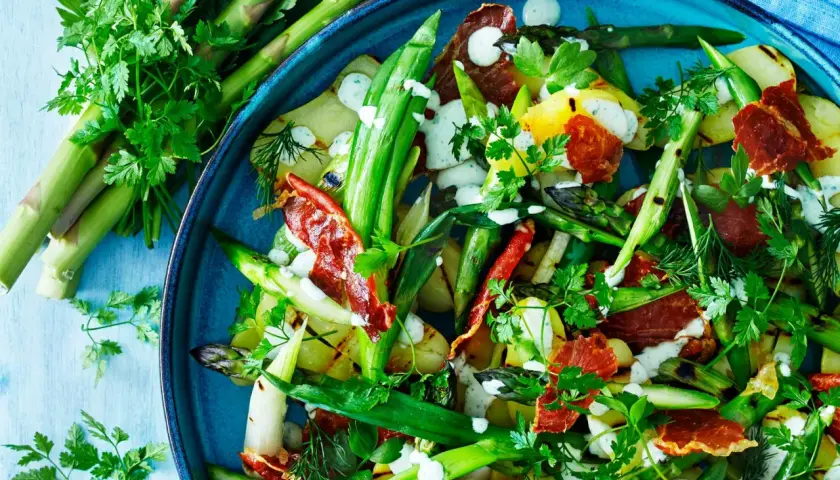 A close-up of a vibrant salad with grilled asparagus, potatoes, and crispy prosciutto, drizzled with a creamy dressing and garnished with fresh herbs.