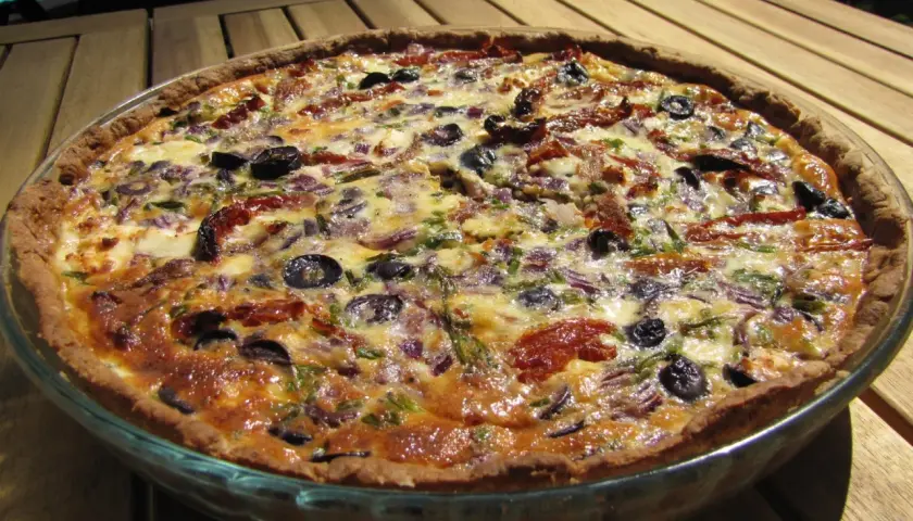 En gyllene paj utan skal, fylld med soltorkade tomater, oliver och lök, serveras på ett träbord.