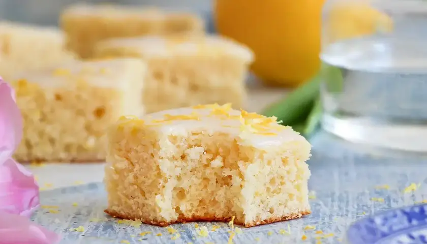 En bit saftig citronkaka i långpanna med glasyr och citronskal. En hel citron och en tulpan syns i bakgrunden.