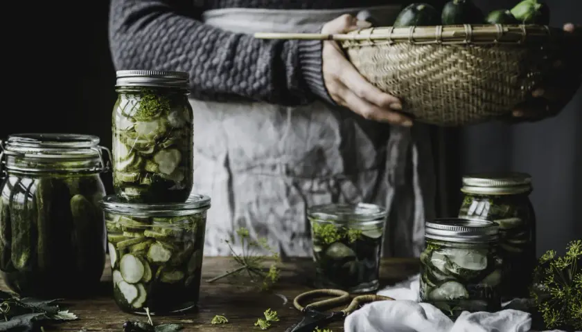 Burkar med inlagd gurka och dill, redo för ett gott saltgurka recept. En korg med färska gurkor i bakgrunden.