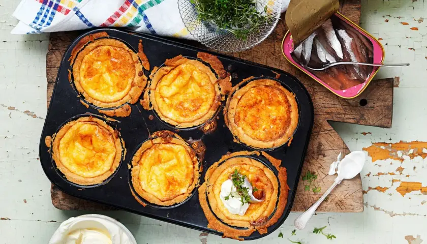Nybakade minipajer med ost i en muffinsform. En burk ansjovis och lite gräddfil står bredvid.