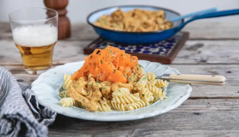 En tallrik med rotfrukter och ost och skinksås över pasta. En öl står bredvid.