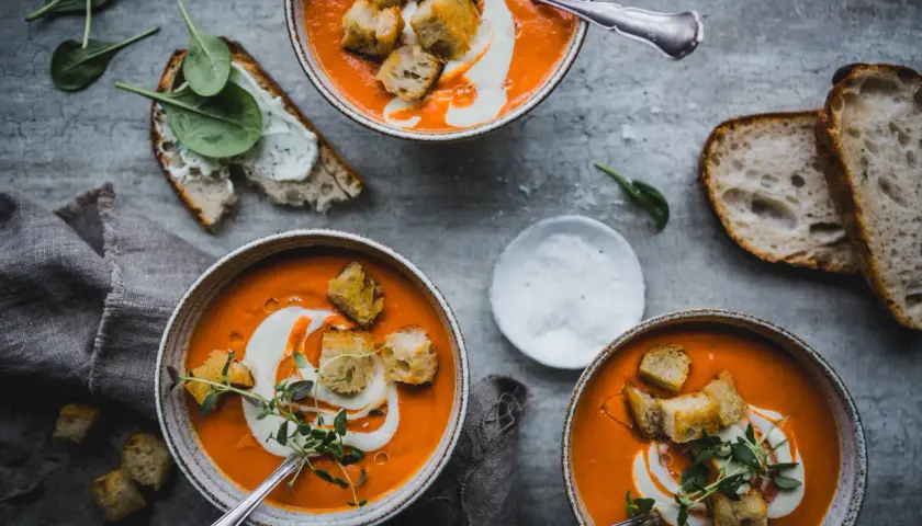 Tre skålar med krämig tomatsoppa, gjord på krossade tomater, toppas med krutonger och en virvel av grädde. Serveras med gott bröd och färsk spenat.