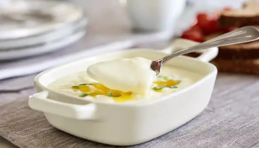 En sked lyfter krämig bechamelsås ur en vit skål, garnerad med gräslök och olja. Perfekt för ditt bechamelsås recept.