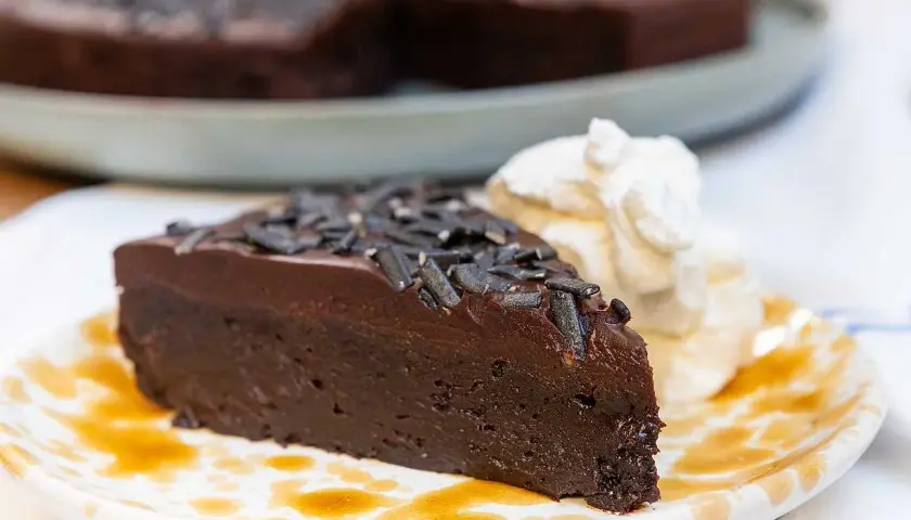 En saftig kladdkaka med lakritsströssel, serverad med vispad grädde och en skvätt sirap på en mönstrad tallrik.