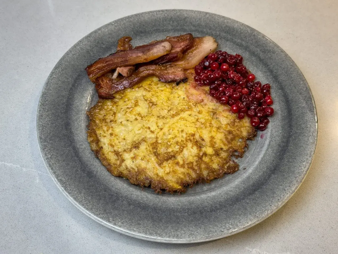 En gyllene raggmunk i ugn serveras med knaperstekt bacon och rårörda lingon på en grå tallrik.