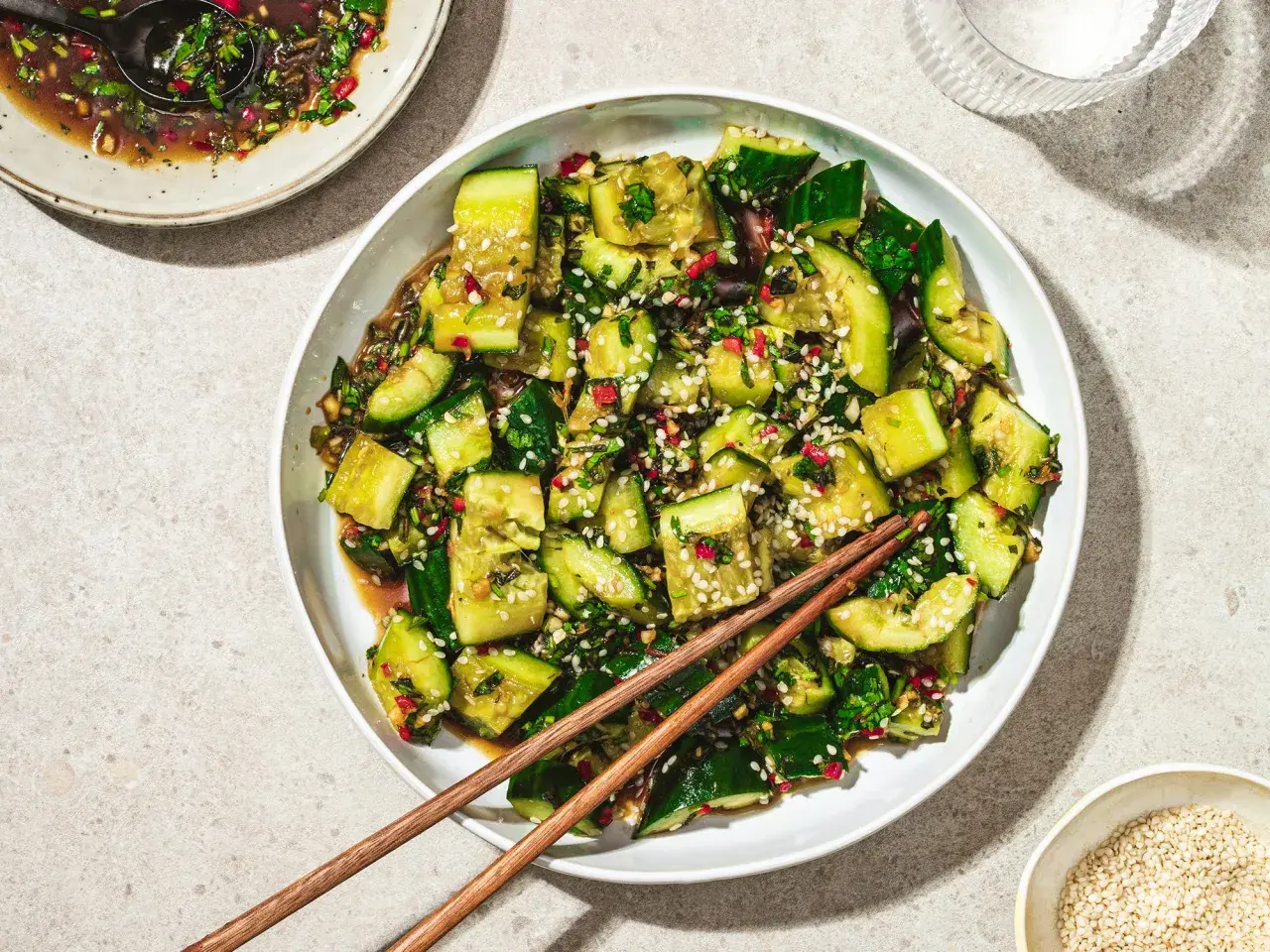 En skål med asiatisk gurksallad, toppad med sesamfrön och färska örter. En skål med dressing och ätpinnar ligger bredvid.