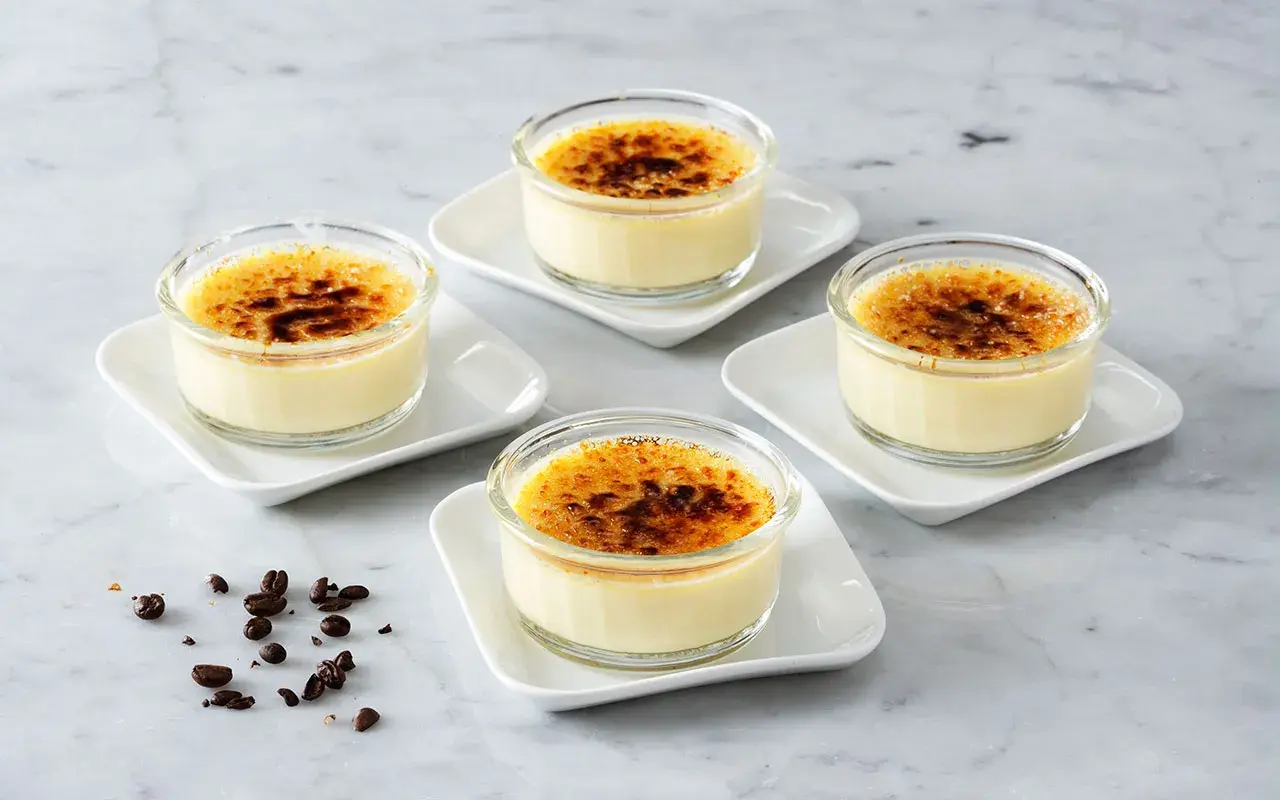 Fyra små glas med krämig creme brulee, toppad med karamelliserat socker. Kaffebönor ligger utspridda på marmorbordet.