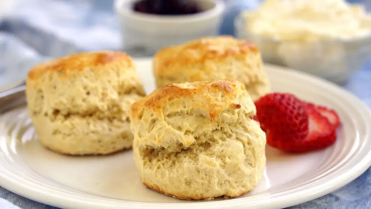 Tre gyllene scones på en vit tallrik, redo att avnjutas med sylt och grädde. Ett perfekt scones recept för fikat.