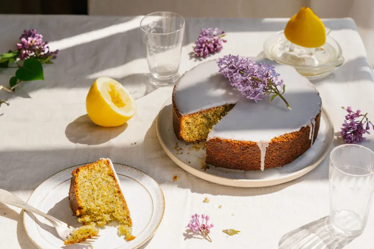 En saftig kaka med citron och vallmofrön, täckt med glasyr och dekorerad med syren. En bit är upplagd på en tallrik med en gaffel.