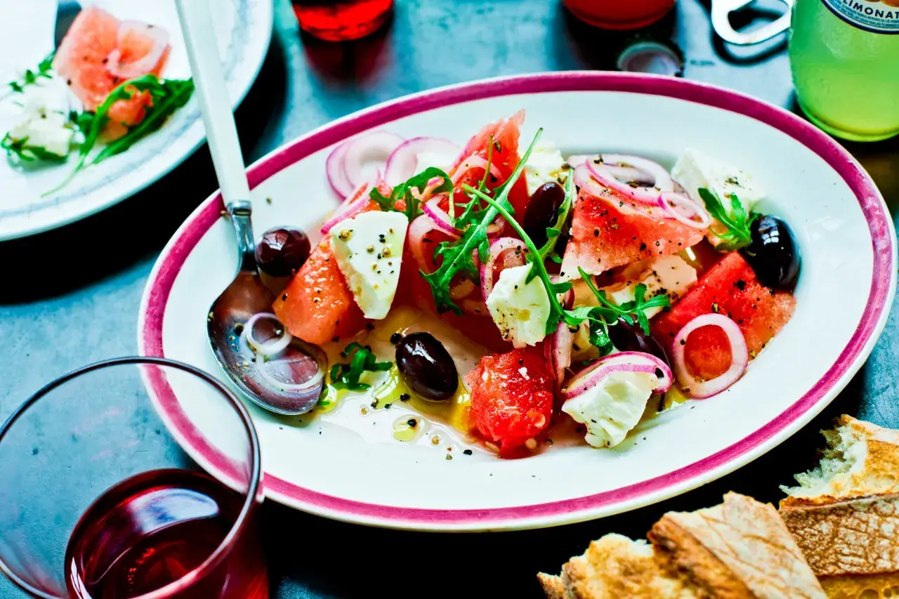 En uppfriskande melon och fetaostsallad med oliver, rödlök och ruccola, serverad med bröd och en drink.