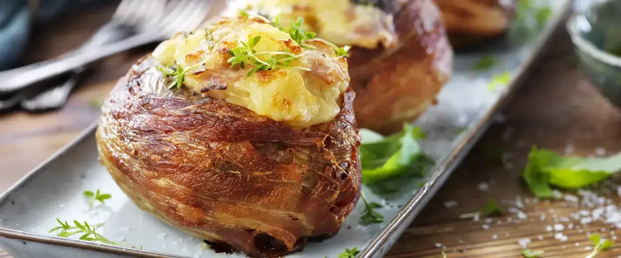 Baked potatoes wrapped in bacon, topped with cheese and herbs, on a platter.