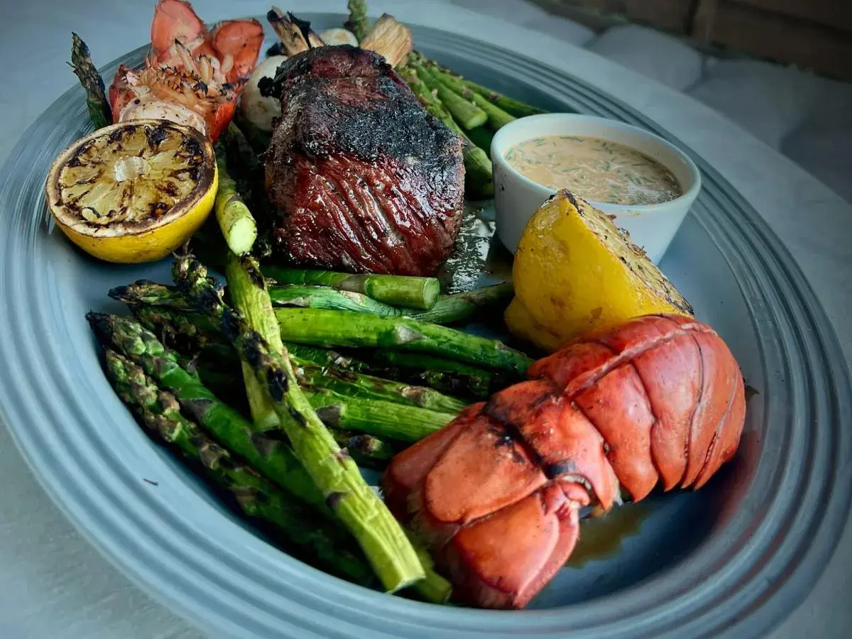 Grilled steak and lobster tail with asparagus, lemon wedges, and a creamy sauce on a platter.