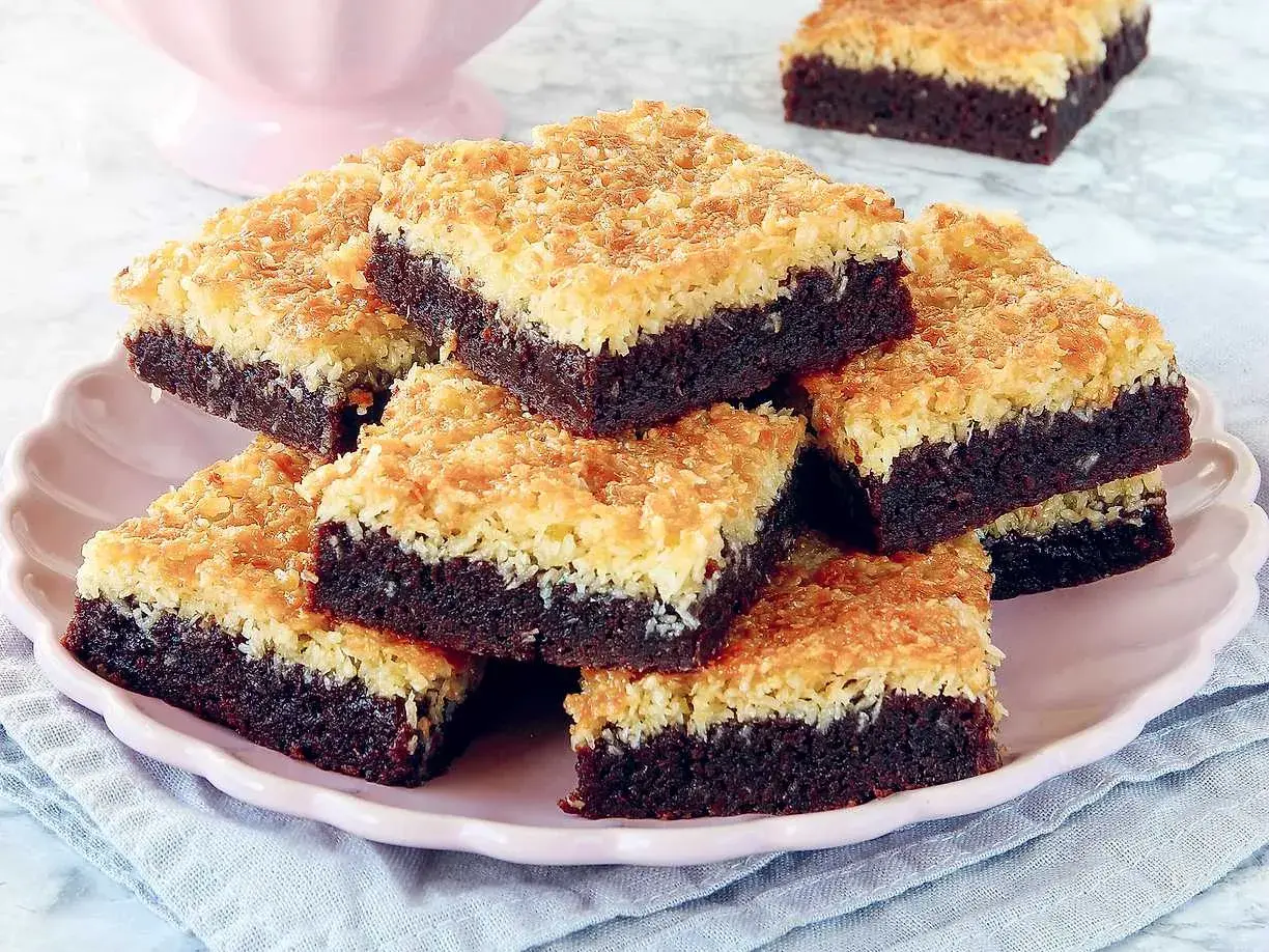 En hög med kladdkaka med kokostosca på en rosa tallrik. Den mörka chokladkakan toppas med en gyllene, krispig kokosbädd.