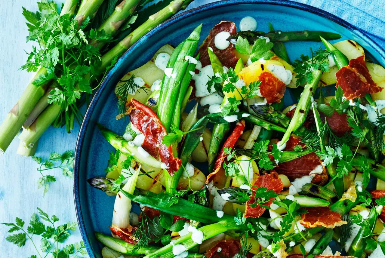 A close-up of a vibrant salad with grilled asparagus, potatoes, and crispy prosciutto, drizzled with a creamy dressing and garnished with fresh herbs.