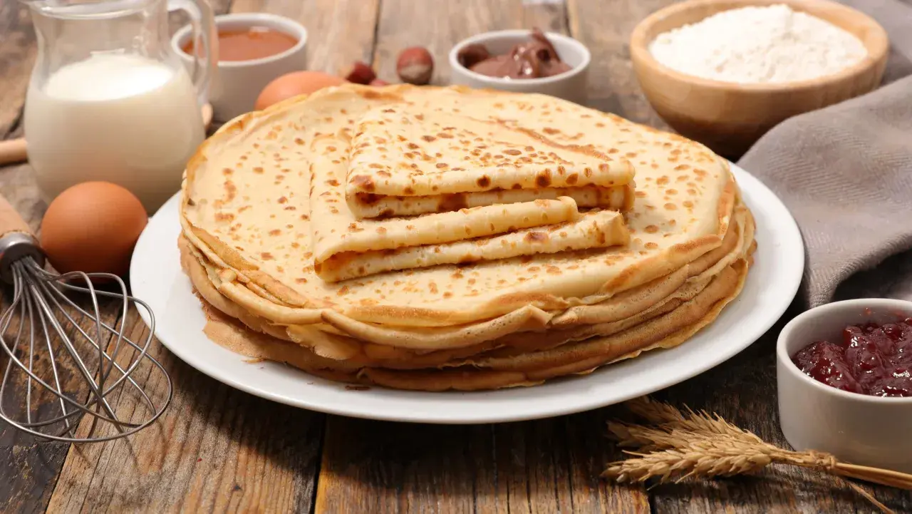 En hög med gyllene pannkakor, redo att avnjutas. Ingredienser som mjölk, ägg och mjöl syns i bakgrunden, perfekt för ett gott pannkakor recept.