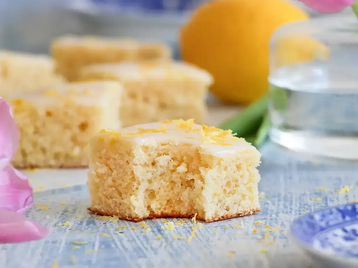 En bit saftig citronkaka i långpanna med glasyr och citronskal. En hel citron och en tulpan syns i bakgrunden.