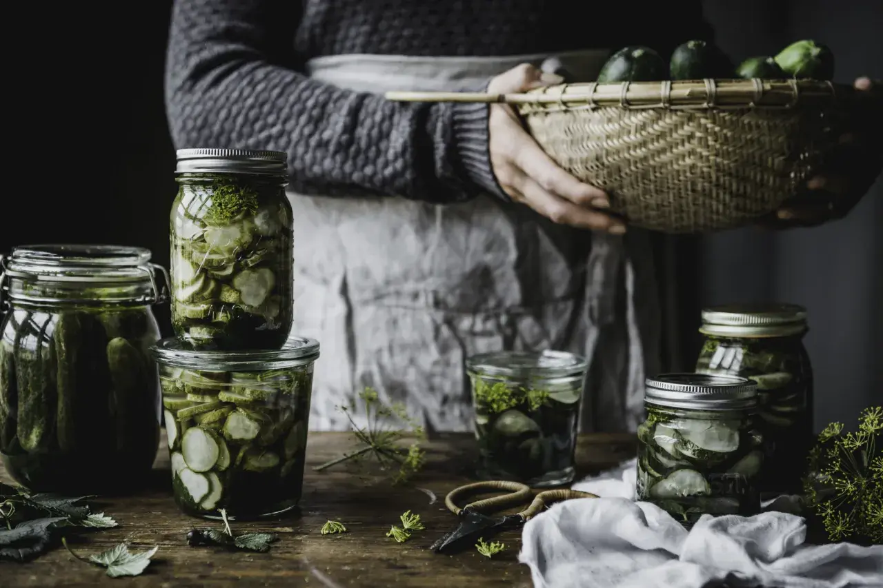 Burkar med inlagd gurka och dill, redo för ett gott saltgurka recept. En korg med färska gurkor i bakgrunden.